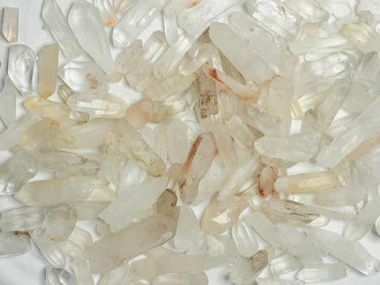A dense overhead arrangement of raw, unpolished clear quartz crystal points on a soft white surface, some with natural earthy orange and rust-colored inclusions, showing the stones in their natural untreated state.